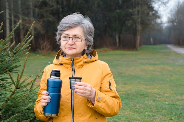 Senior woman walking outside in the forest. Elderly woman holding thermos and mug of hot tea. Concept hike, warming drink in cold weather.