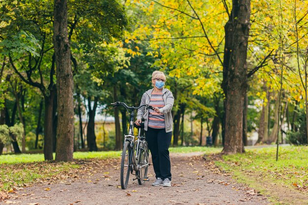 Donna senior cammina nel parco con una bicicletta in una maschera medica protettiva