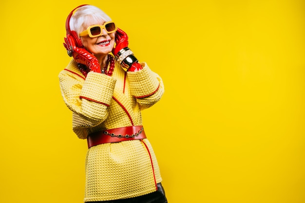 Senior woman wearing hat listening music against yellow background