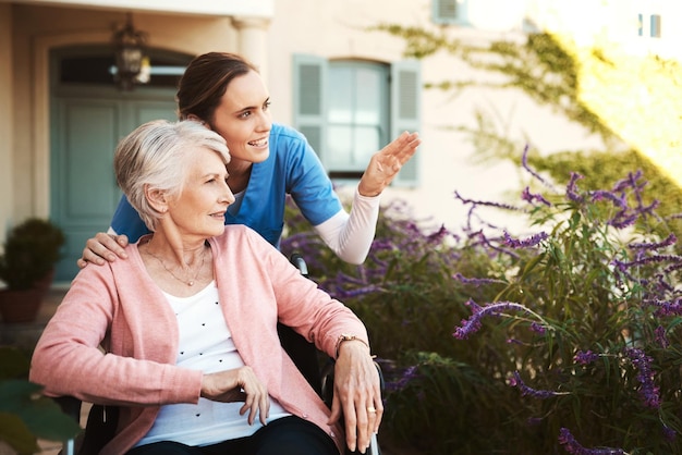 Senior woman wheelchair and nurse in garden pointing and talking on walk in healthcare support at nursing home Happy elderly female or caregiver showing patient or person with a disability outdoors