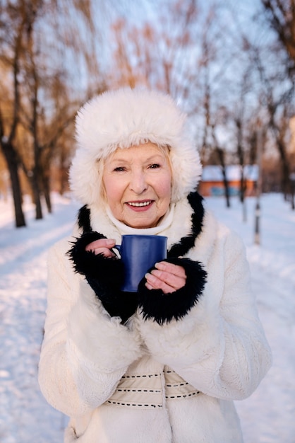 雪の森で手に青いカップを保持している白い帽子と毛皮のコートの年配の女性