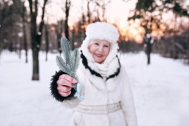 雪の森で手にトウヒの枝を保持している白い帽子と毛皮のコートの年配の女性