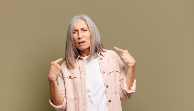 Senior donna con un cattivo atteggiamento che sembra orgogliosa e aggressiva, rivolta verso l'alto o facendo segno divertente con le mani