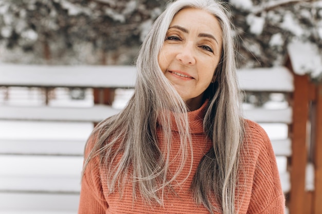 Donna anziana con i capelli grigi in un maglione lavorato a maglia che sorride sinceramente su uno sfondo invernale