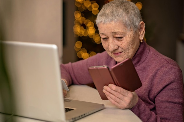 Senior woman with gray hair sits in front of a laptop with a phone in her hands, enters card detail