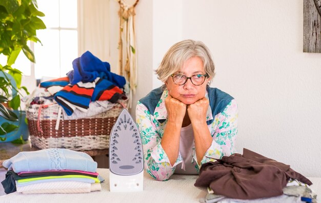 A senior woman with grey hair sad because too much clothes to iron, domestic room with plants and white wall.