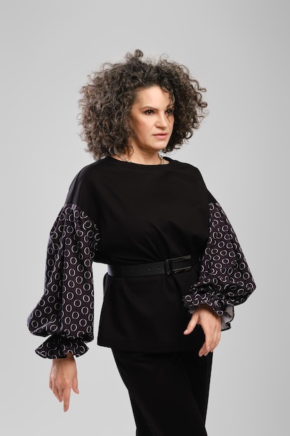 Senior woman with lush curly hair and black pantsuit posing in studio over grey background