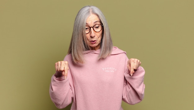 Senior woman with open mouth pointing downwards with both hands, looking shocked, amazed and surprised