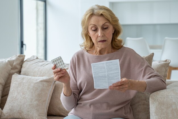 La donna anziana con le pillole legge le istruzioni a casa seduta sul divano