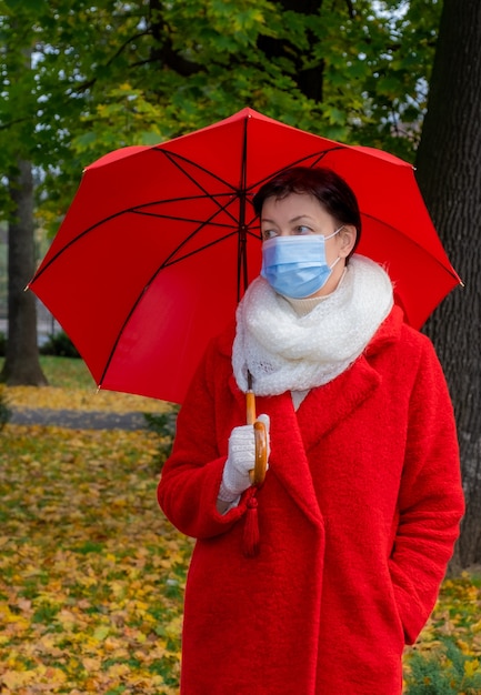 Senior woman with protective medical mask on her face walks in autumn park with red umbrella.