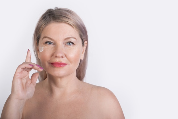 Senior woman with undressed shoulders is posing on a white studio wall advertising something while applying facial cream near eyes