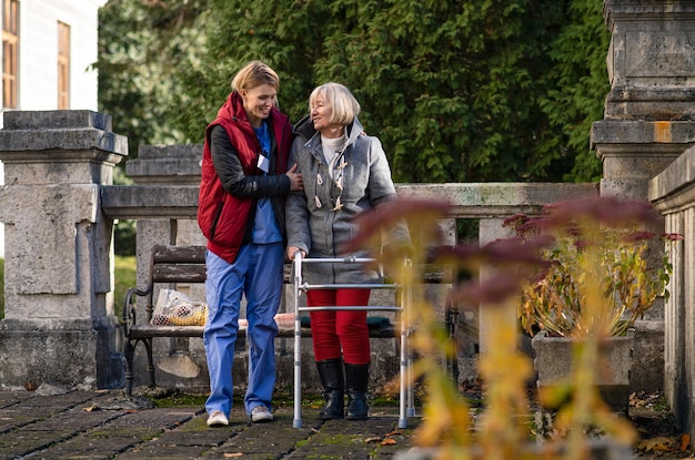 Senior woman with walking frame and caregiver or healthcare worker outdoors on a walk in park.