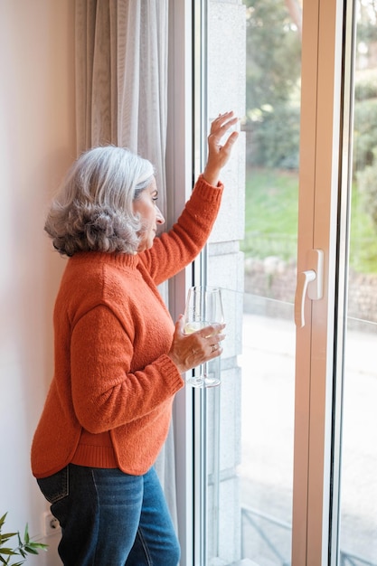 Senior woman with white hair having a glass of wine while relaxing looking out the window at home Concept Lifestyle relax disconnection