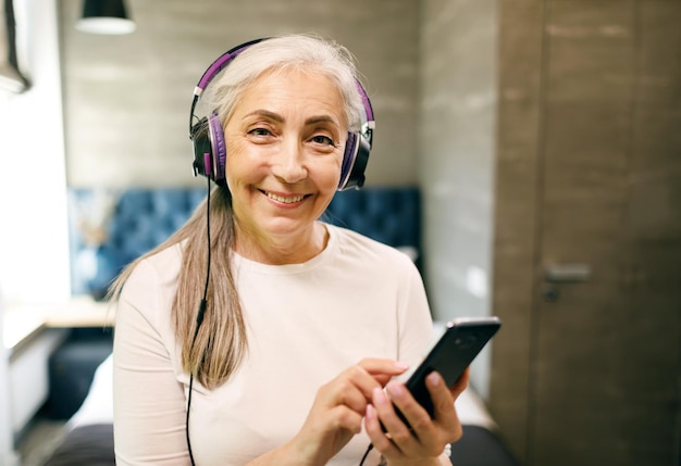 Senior woman with white hair in headphones searching her favorite songs on the smartphone and smiling to the camera