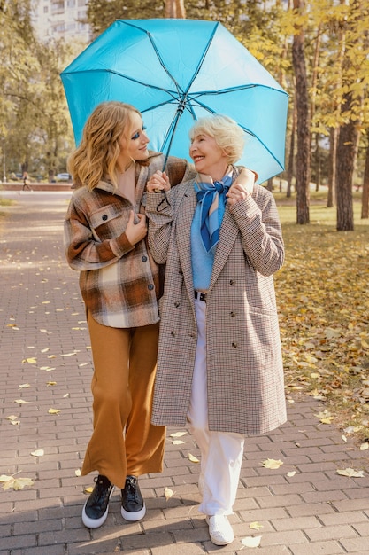 senior woman with young daughter walking outdoor under blue umbrella in autumn park