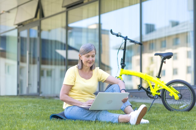 Senior woman working on laptop remotely sitting on the grass in the park