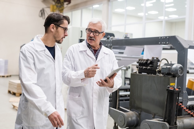 Esperti di qualità senior e giovani in camici da laboratorio utilizzando tablet durante il controllo delle apparecchiature di stampa presso l'impianto di stampa