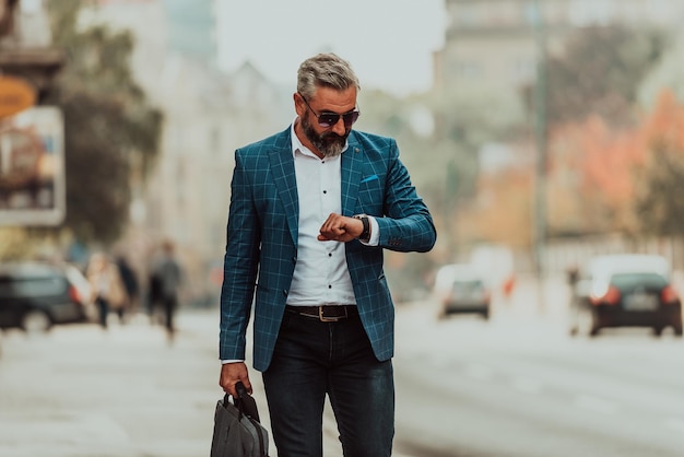 Senior zakenman die door de stad loopt, een tas draagt en een horloge gebruikt.