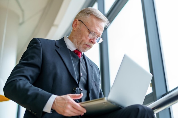 Senior zakenman in de buurt van het panoramische raam Spreken met collega's via de laptop Videoconferentie Werk op afstand Isolatie en quarantaineconcept