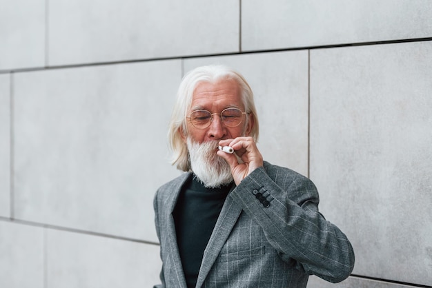 Senior zakenman in formele kleding met grijs haar en baard rookt buiten elektronische sigaret