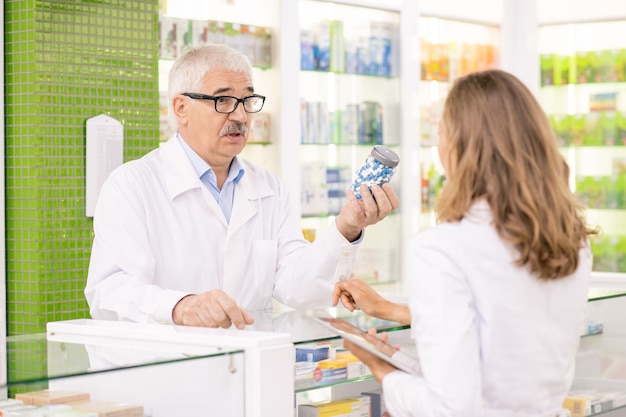 Senior zelfverzekerde apotheker in witte jas en bril die zijn jonge assistent-fles pillen laat zien terwijl hij de kenmerken ervan beschrijft