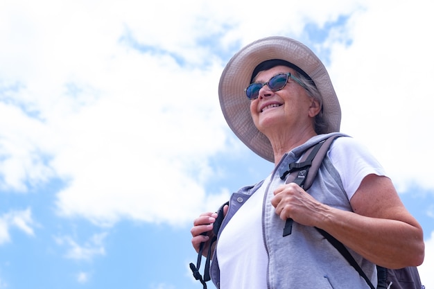 Senior zorgeloze vrouw met rugzak en pet genieten van buiten excursie. Blauwe lucht