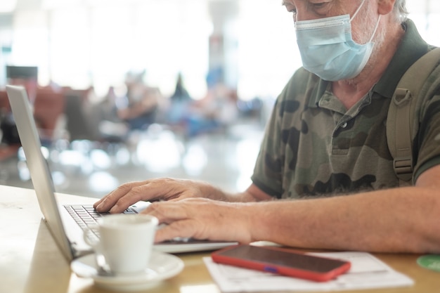 Seniour man with face mask drinks a coffee and uses his laptop in the airport waiting for boarding