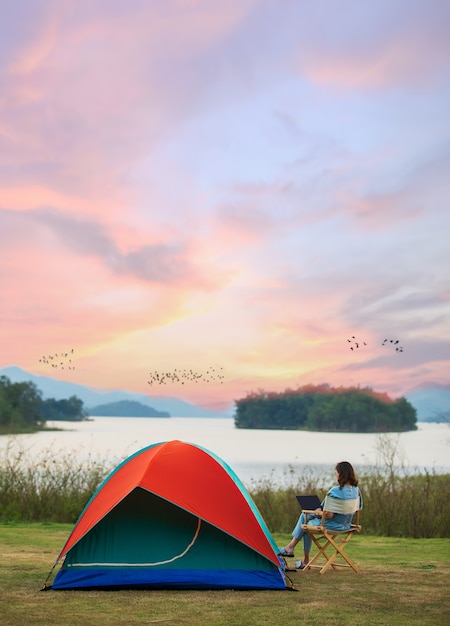 A sense of peace and serenity photo of female traveler sitting beside camping tent and using notebook laptop computer working from the lake side. There are group of birds flying in colorful sky.