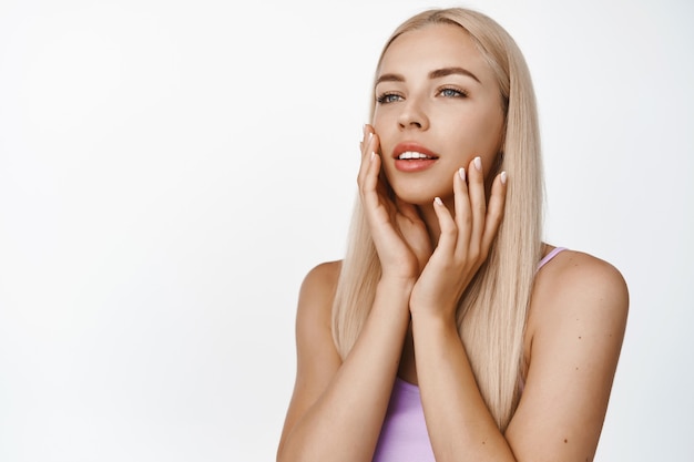 Sensual blond woman touching her soft glowing skin on face, gazing away, concept of cosmetology and beauty, standing on white.