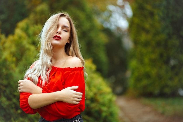 Sensual blonde girl in red blouse posing with naked shoulder at the thuja trees. Space for text