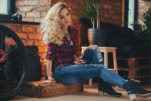 Sensual blonde hipster girl with long curly hair dressed in a fleece shirt and jeans holds a smartphone sitting on a wooden box at a studio with loft interior.