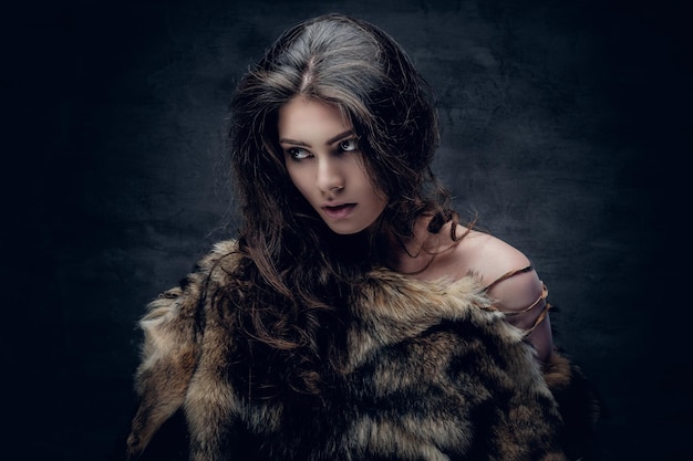 Sensual brunette female with curly hair dressed in a fur coat posing in deep shadow on grey background in a studio.