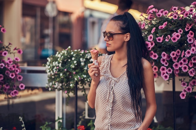 トレンディな服を着てサングラスをかけた官能的なブルネットの女の子は、花で飾られたテラスの近くに立っている間、夏の日を楽しんでいます。ストロベリーアイスクリームを持っています。