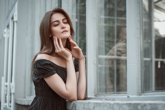 Sensual brunette girl wearing black dress with naked shoulders, posing at the greenhouse. Empty space