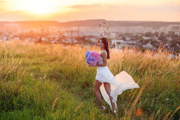 夕暮れ時の芝生の中を柔らかい色の花の花束と光の白い夏のドレスで官能的なかわいい女の子が歩きます。