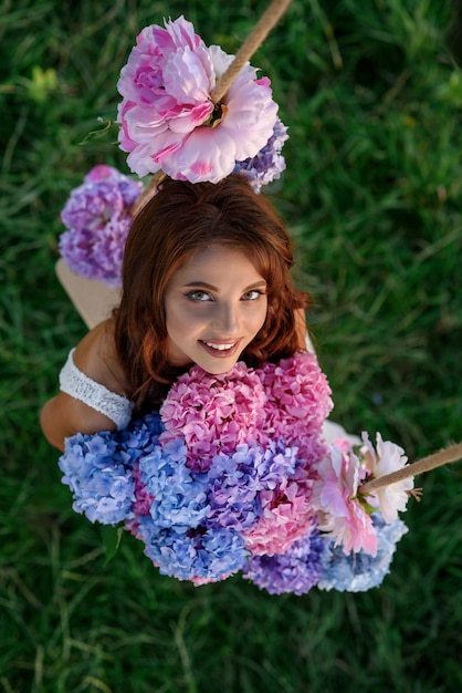 Foto ragazza sveglia sensuale con il sorriso perfetto in un vestito bianco che si siede su un'oscillazione di legno con un mazzo di fiori colorati teneri, vista superiore.