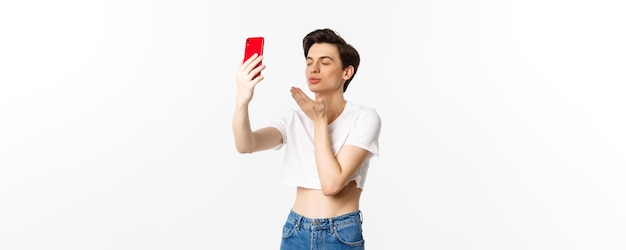 Sensuale uomo gay in crop top che si fa selfie sullo smartphone inviando un bacio d'aria alla fotocamera del telefono e chiudendo e