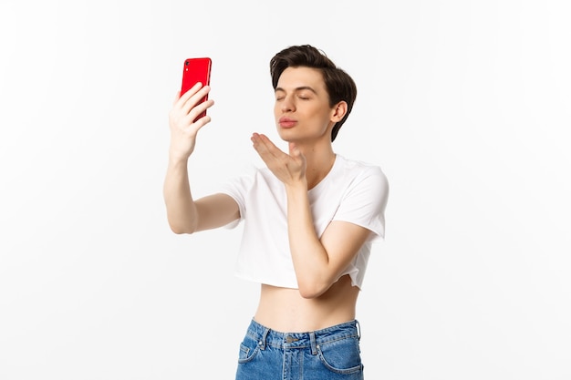 Sensual gay man in crop top taking selfie on smartphone, sending air kiss at phone camera and close eyes dreamy, standing over white background.