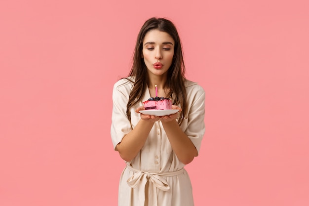 Sensuale, bellissima ragazza bruna caucasica di b-day festeggia il suo compleanno, tenendo il piatto di torta b-day e spegnendo la candela, facendo desiderio e sorridendo con gioia, in piedi rosa spensierato