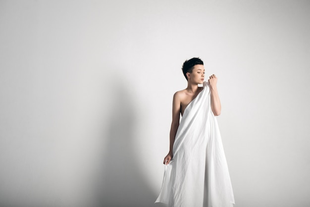 Sensual naked young adult caucasian woman, wrapped in a satin, silk sheet against grey background. High contrast