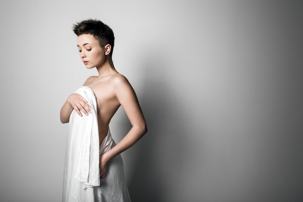 Sensual naked young adult caucasian woman, wrapped in a satin, silk sheet against grey background. High contrast