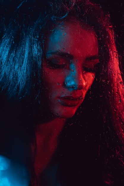 Ritratto sensuale di bella ragazza dietro vetro con le gocce di pioggia con illuminazione blu rossa