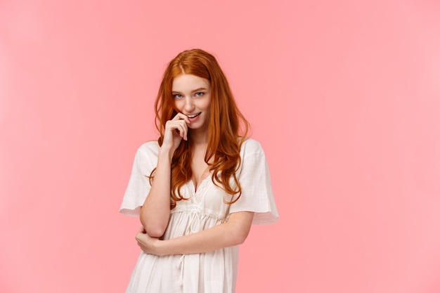 Sensual, romantic and sassy, seductive redhead daring woman in white dress, biting finger and coquettish look  from under forehead, standing pink wall, flirting