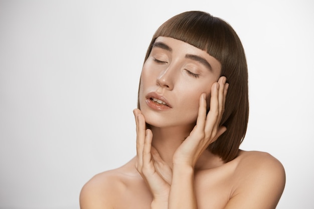 Foto donna sensuale con pelle fresca perfetta e capelli lucenti, bella donna con trucco nudo e acconciatura alla moda con frangia che tocca il viso con gli occhi chiusi