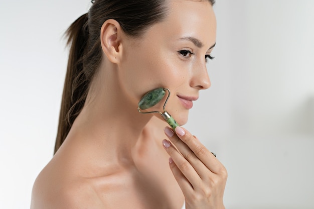 Sensual young woman massaging face with jade roller over white\
studio background. caucasian model taking care of natural beauty.\
beauty routine and skin procedures concept.