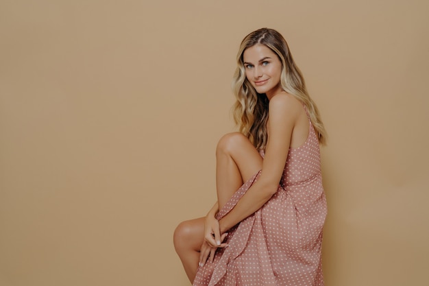 Sensual young woman in summer dress sitting sideways on chair over beige wall studio background with copy space, Beautiful charming female with long blonde hair posing with romantically tender smile