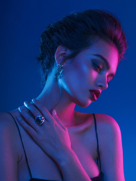 Sensual young woman with bare shoulders, hairstyle and makeup, wear in grandiose blue stone jewelry, over dark purple neon lights.