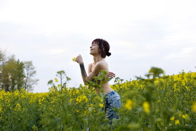 Sensual young woman with tattoos posing in a rapeseed field\
among yellow flowers on a sunny day.