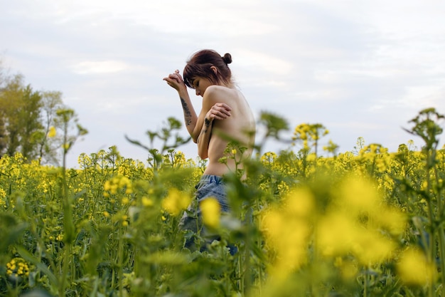 Sensual young woman with tattoos posing in a rapeseed field\
among yellow flowers on a sunny day.