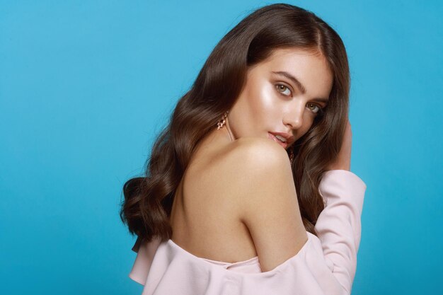 Sensual young woman with wavy brunette hairstyle and makeup, dressed in pink blouse whith bare shoulder, isolated blue background.